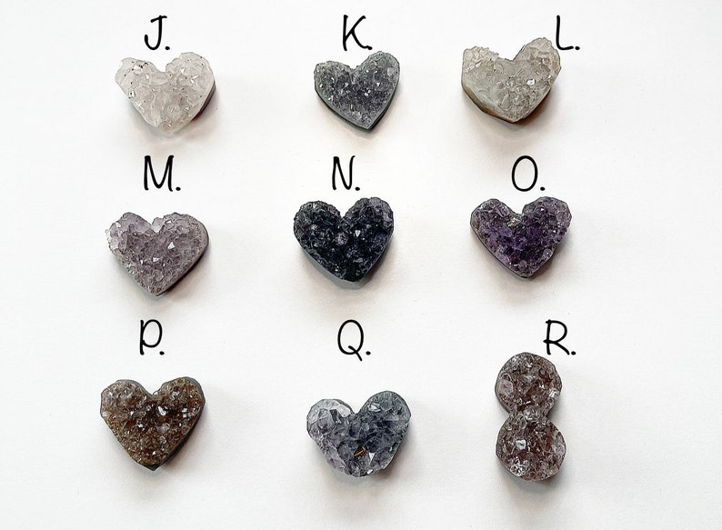 Puede incluir: Nueve cristales de amatista en forma de coraz&oacute;n, etiquetados de la J a la R, en varios tonos de morado, gris y blanco.