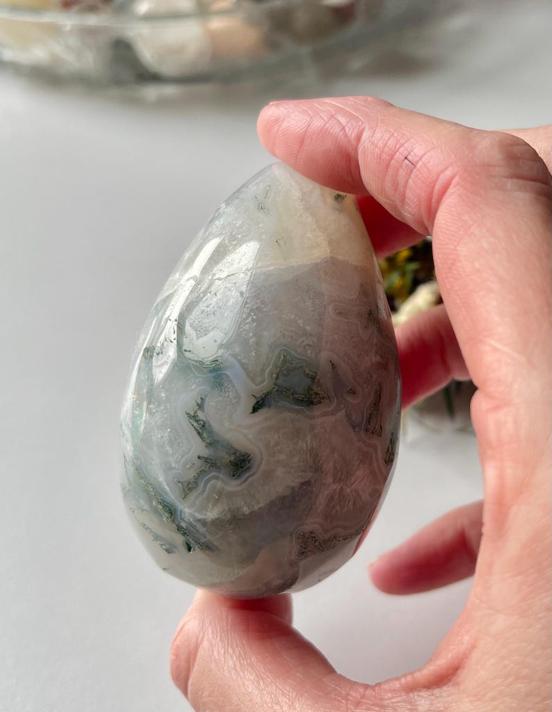 May include: A polished, egg-shaped moss agate stone featuring translucent white and grey areas with intricate, dark green dendritic inclusions that resemble fern-like patterns throughout the smooth, rounded surface of the mineral specimen.