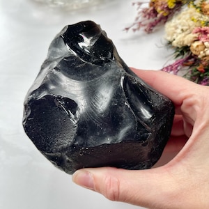 May include: A large, irregular chunk of black obsidian stone with a smooth, glossy, and reflective surface. The rock features sharp, conchoidal fracture patterns and edges, displaying a deep, solid black colour throughout its entire structure.