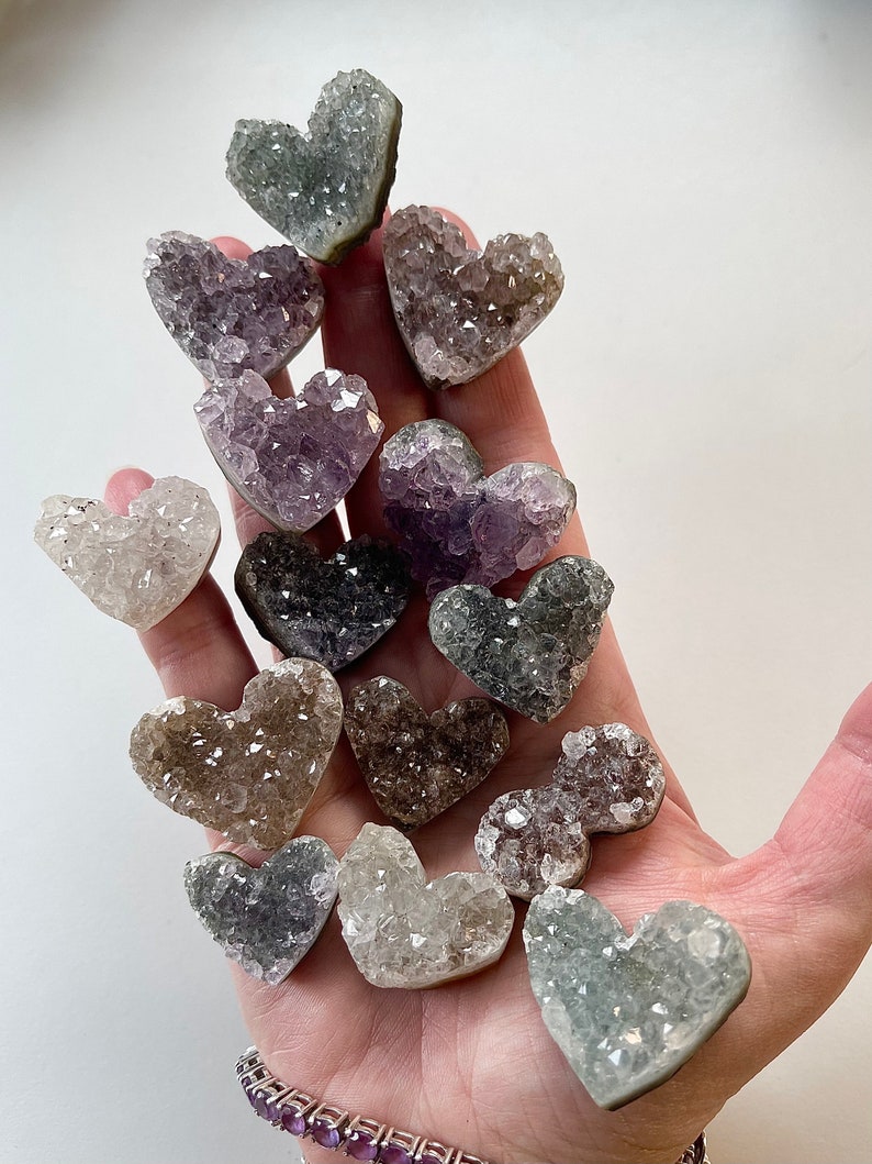 Puede incluir: Una colecci&oacute;n de cristales de amatista en forma de coraz&oacute;n en varios tonos de morado, gris y blanco. Los cristales est&aacute;n dispuestos en una pila sobre una mano.