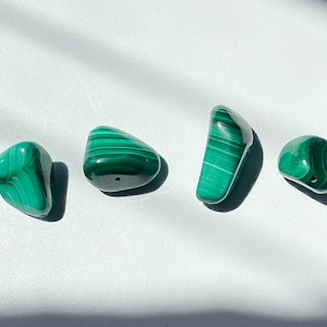 May include: Seven smooth, green malachite stones with a variety of shapes and sizes. The stones are arranged in a row on a white surface.