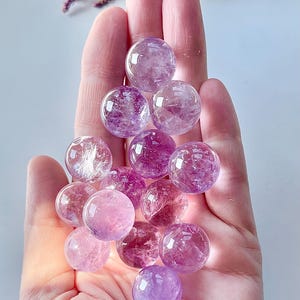 May include: A collection of small, round, amethyst crystal spheres in various shades of purple and pink, held in a hand.