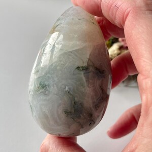 May include: A polished, teardrop-shaped gemstone with a mix of white, gray, and green hues. The stone has a translucent quality, revealing internal patterns and inclusions. The smooth surface reflects light, highlighting its natural beauty.