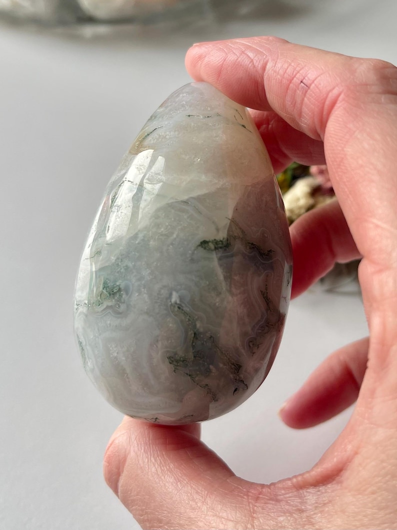 May include: A polished, teardrop-shaped gemstone with a mix of white, gray, and green hues. The stone has a translucent quality, revealing internal patterns and inclusions. The smooth surface reflects light, highlighting its natural beauty.