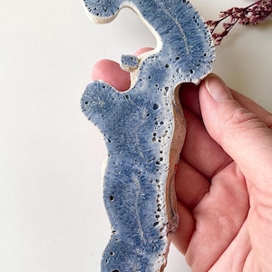 May include: A blue and beige ceramic sculpture with a unique, organic shape. The sculpture has a textured surface with a porous appearance and is held by a hand. The blue color is concentrated in the center, with beige edges.