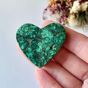 May include: A heart-shaped malachite stone with a swirling pattern of dark and light green. The polished surface reveals intricate details, creating a unique and natural design. The stone is held in a hand.