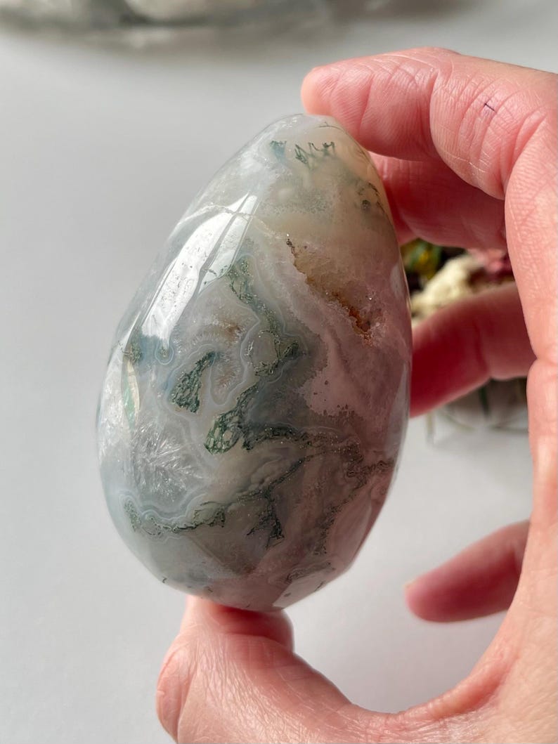 May include: A polished, egg-shaped moss agate stone featuring translucent white and gray areas with intricate, dark green dendritic inclusions that resemble branching plant-like patterns throughout the smooth, rounded surface.