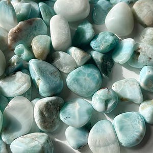 May include: A close-up of a collection of polished larimar stones. The stones are in shades of blue, white, and brown. The smooth, rounded stones are arranged closely together, creating a textured surface. The stones are of various sizes and shapes.