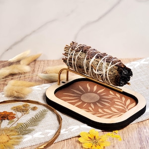 May include: A sage smudge stick sits on a wooden tray featuring a sun and leaf design. The tray has a black border and a gold-coloured metal holder. Dried flowers and decorative elements surround the tray, creating a natural, earthy aesthetic.