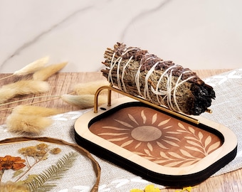 May include: A sage smudge stick rests on a wooden tray with a sun and leaf design. The tray has a black border and a gold-colored metal holder. Dried flowers and decorative elements surround the tray, creating a natural, earthy aesthetic.