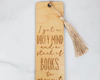 May include: Wooden bookmark with the engraved text "I got a DIRTY MIND and a stack of BOOKS to prove it." The bookmark has a tassel attached and a hole at the top. The bookmark is made of light-colored wood.