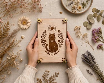 May include: A rectangular light-colored wooden flower press featuring a dark brown laser-engraved illustration of a seated cat centered within a floral wreath and a sun symbol above. The press is secured at each corner with brass-colored metal hardware.