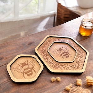 Puede incluir: Posavasos hexagonales de madera con un diseño de abeja grabado con láser. Los posavasos son de color marrón claro con un borde marrón oscuro. Un posavasos es más pequeño que el otro. El más grande tiene un borde floral.