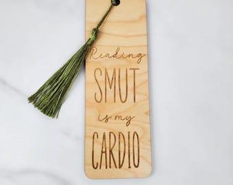 May include: Wooden bookmark with the phrase "Reading SMUT is my CARDIO" engraved in a playful font. The bookmark has a green tassel attached at the top. The wood has a light, natural finish.