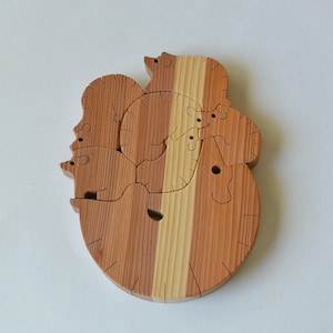 Può includere: Un puzzle di legno con diverse forme di animali, tra cui un orso, un cervo e un uccello. I pezzi del puzzle sono in legno naturale di colore marrone chiaro e marrone rossiccio.