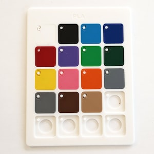 May include: A white plastic color chart with the word "PLA" at the top. The chart has 15 squares, each with a different color of plastic. The colors include white, black, blue, dark blue, red, purple, green, dark green, yellow, pink, orange, gray, brown, tan, and a blank square.