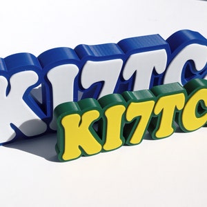 May include: Three dimensional letters spelling "KITCJ" in blue, white, green, and yellow. The letters are stacked on top of each other.