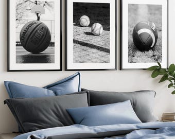 May include: A set of three black and white framed wall art prints. The left print shows a Spalding basketball on a court. The middle print features two baseballs on a textured surface. The right print displays an American football on grass.