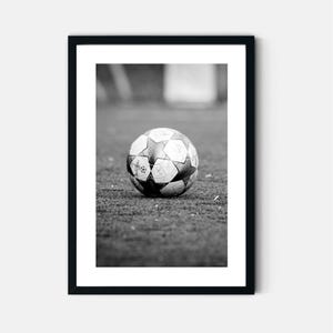 May include: A black and white framed wall art print featuring a soccer ball with a star-patterned design resting on a field. The ball displays the UEFA Champions League logo and text. The artwork is enclosed in a simple black rectangular frame.