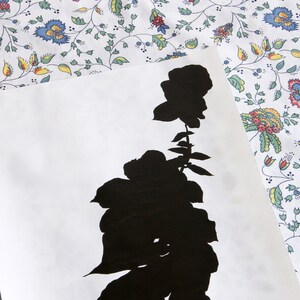 May include: A black silhouette of a rose with leaves and stem on a white background. The floral design is set against a patterned fabric with a white background and colorful flowers.