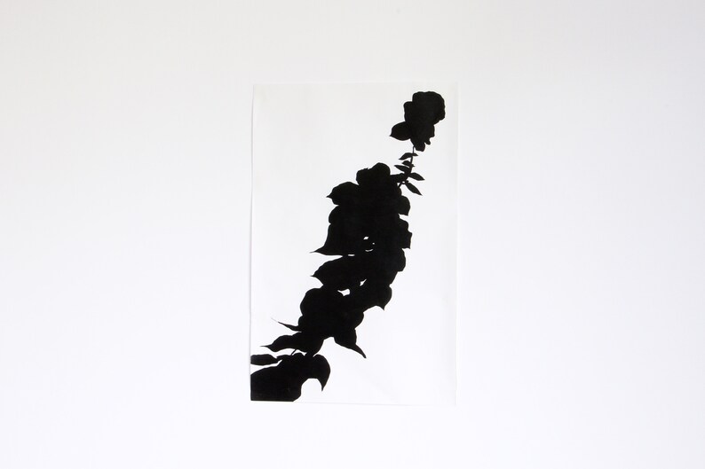 May include: A black floral silhouette print on a white rectangular paper. The artwork features a stylized depiction of a flower stem with leaves and blooms. The contrast between the black design and the white background creates a striking visual.