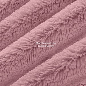May include: Close-up of a soft, plush fabric in a dusty rose color. The fabric has a fuzzy texture and is arranged in diagonal lines. The text "Misty Mauve Luxe Cuddle Bunny" is printed on the fabric.