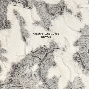 May include: Close-up of a soft, plush fabric in a gray and white color scheme. The fabric has a textured, fluffy appearance, resembling the fur of a baby calf. The text "Graphite Luxe Cuddle Baby Calf" is printed on the fabric.