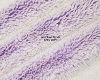 May include: Close-up of a soft, plush fabric with a gradient of white and purple. The fabric has a fuzzy texture and is ideal for making cuddly toys. The text "Aster Luxe Cuddle Snow Bunny" is printed on the fabric.