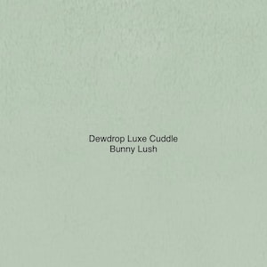 May include: A light green background with the text "Dewdrop Luxe Cuddle Bunny Lush" in a simple, sans-serif font. The background has a textured appearance, resembling fabric or a soft surface.