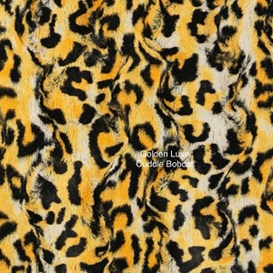 May include: A close-up of a plush fabric with a golden yellow and black leopard print. The fabric is soft and fuzzy, and the print is realistic. The text "Golden Luxe Cuddle Bobcat" is printed on the fabric.