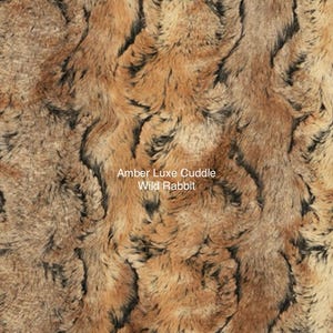 May include: Close-up of a faux fur fabric with a brown, black, and beige color scheme. The fabric has a soft, plush texture and a wild rabbit pattern. The text "Amber Luxe Cuddle Wild Rabbit" is printed on the fabric.