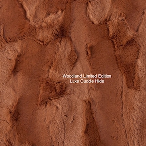 May include: Close-up of a brown faux fur fabric with a soft, plush texture. The fabric is labeled "Woodland Limited Edition Luxe Cuddle Hide."