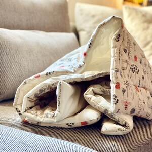 May include: A white and cream colored quilt with a floral and insect print. The quilt is folded and sitting on a gray couch.