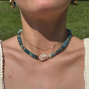 Op de afbeelding: Een ketting met turquoise, blauwe en bruine kralen, met een grote, onregelmatig gevormde witte parel hanger. Een fijne gouden ketting is ook zichtbaar. De ketting wordt gedragen over een crèmekleurige top.