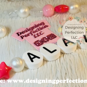 Puede incluir: Un collar de nombre para ni&ntilde;a de color rosa y blanco con el nombre "Alana" escrito en cuentas blancas con letras negras. El collar est&aacute; hecho con cuentas rosas y blancas y estrellas. El collar est&aacute; sobre una superficie blanca y peluda. El texto "Designing Perfection LLC" es visible en la imagen.