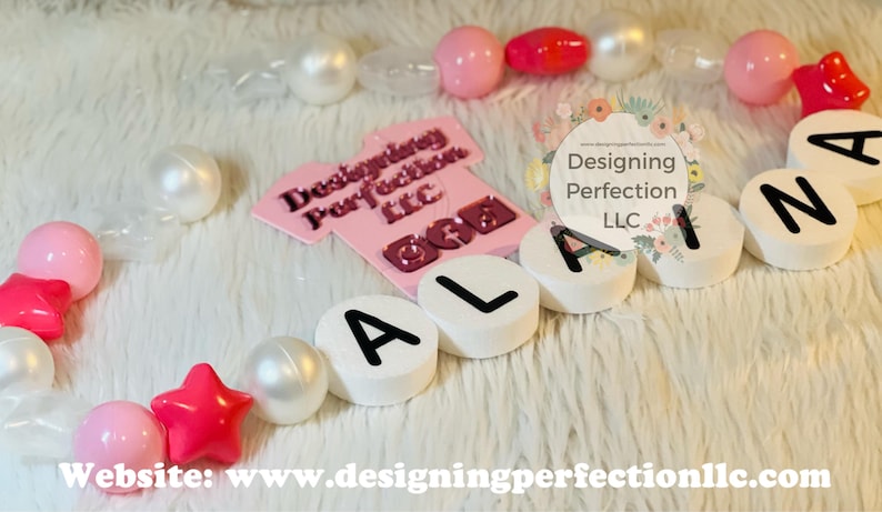 Puede incluir: Un collar de nombre para ni&ntilde;a de color rosa y blanco con el nombre "Alana" escrito en cuentas blancas con letras negras. El collar est&aacute; hecho con cuentas rosas y blancas y estrellas. El collar est&aacute; sobre una superficie blanca y peluda. El texto "Designing Perfection LLC" es visible en la imagen.