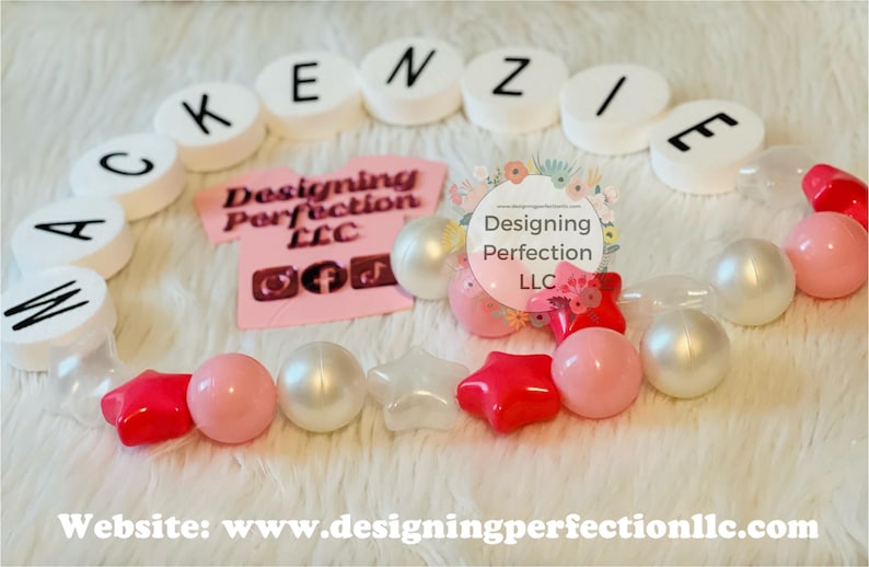 Puede incluir: Un collar de cuentas blancas y rosadas con cuentas de letras blancas que deletrean el nombre "MACKENZIE". El collar tambi&eacute;n presenta cuentas en forma de estrella blancas y rosadas. El collar est&aacute; sobre una superficie blanca y peluda. El texto "Designing Perfection LLC" es visible en la imagen.