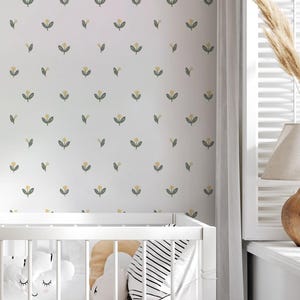 Op de afbeelding: Een witte wieg met een bloemenbehang achtergrond. De wieg is voorzien van wit beddengoed, kussens en een deken. Een rond, beige tapijt ligt op de houten vloer. Een geweven mand bevat witte handdoeken. De kamer is ingericht in neutrale tinten.