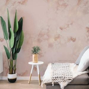Può includere: Un muro rosa chiaro con un effetto acquerello, che presenta un tavolino bianco con un vaso dorato e una pianta verde. Un divano grigio con una coperta bianca e cuscini è in primo piano. Il testo 'WEST COAST WALLS' è visibile sul muro.