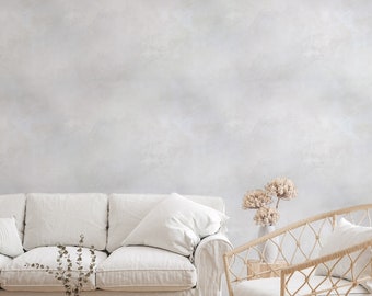 May include: A white sofa with a white throw pillow and a wicker chair with a white cushion. The sofa and chair are in front of a light gray wall. There is a small round woven ottoman in front of the sofa. A small vase with dried flowers is on a wooden table next to the wicker chair.