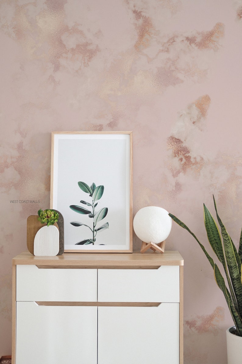 Può includere: Una cassettiera bianca con due cassetti e due ante, una stampa incorniciata di una pianta verde, una lampada lunare bianca e una pianta di serpente in vaso davanti a una parete testurizzata rosa e oro. Il testo 'WEST COAST WALLS' è visibile nell'angolo inferiore sinistro dell'immagine.
