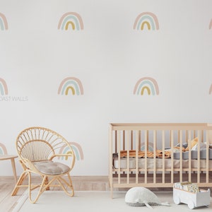 May include: A nursery room with a white wall decorated with multiple pastel rainbow decals. A wooden crib with a white mattress and a gray blanket is in the center of the room. A wicker chair with a gray cushion is in the foreground. The text "WEST COAST WALLS" is visible on the wall.