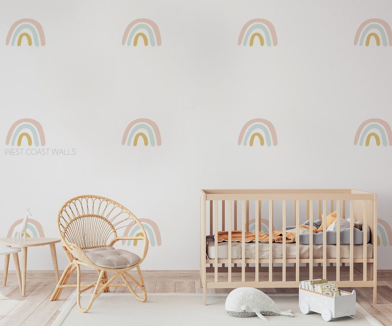 May include: A nursery room with a white wall decorated with multiple pastel rainbow decals. A wooden crib with a white mattress and a gray blanket is in the center of the room. A wicker chair with a gray cushion is in the foreground. The text "WEST COAST WALLS" is visible on the wall.