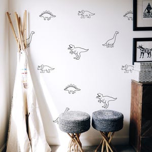 May include: A white wall adorned with black dinosaur decals. Two grey woven stools with wooden legs are on a light grey carpet. A wooden teepee frame with a white canvas cover is in the corner.