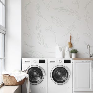 Op de afbeelding: Een lichte, moderne wasruimte met twee witte wasmachines. Een gevlochten mand bevat opgevouwen witte handdoeken. De muren zijn wit met een subtiel bloemmotief. Een kleine plant en schoonmaakmiddelen staan op het aanrecht.
