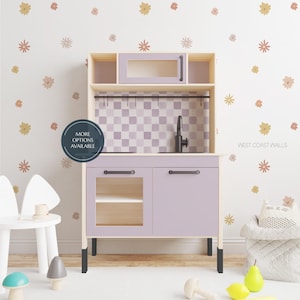 May include: A wooden toy kitchen in light purple with a checkered backsplash. It has a microwave, sink, and storage. The text "MORE OPTIONS AVAILABLE" is on a circular sticker. The wall has floral decals and the text "WEST COAST WALLS".