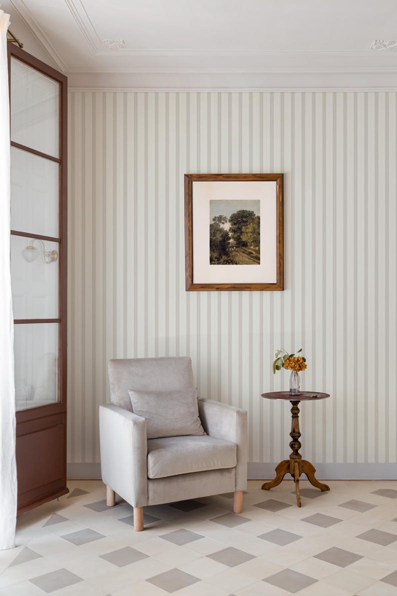 May include: A gray armchair with a pillow sits in a room with white and gray striped wallpaper. A small wooden table with a vase of flowers sits next to the chair. The floor is tiled in a checkered pattern.
