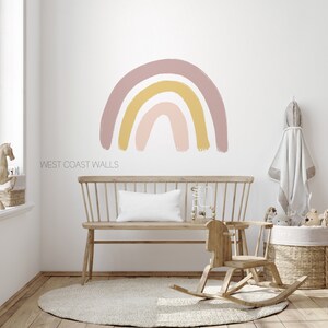 May include: A nursery room with a light pink, yellow, and white rainbow wall decal. The room features a wooden rocking horse, a wooden bench with a white pillow, and a woven basket.
