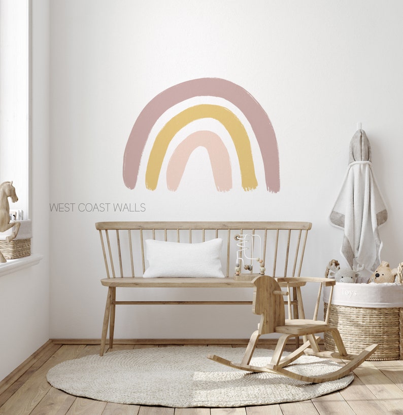May include: A nursery room with a light pink, yellow, and white rainbow wall decal. The room features a wooden rocking horse, a wooden bench with a white pillow, and a woven basket.