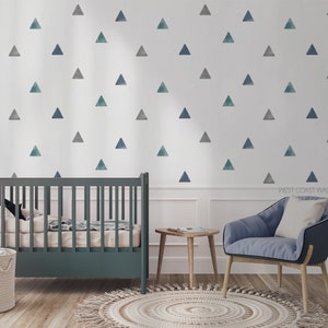 Könnte beinhalten: Ein Kinderzimmer mit einer weißen Wand, die mit blauen und grauen Aquarell-Dreiecksaufklebern bedeckt ist. Ein Kinderbett mit einem dunkelgrauen Holzrahmen steht im Raum, zusammen mit einem blauen Sessel und einem kleinen Holz-Beistelltisch. Der Text "WEST COAST WALLS" ist in der rechten unteren Ecke des Bildes sichtbar.